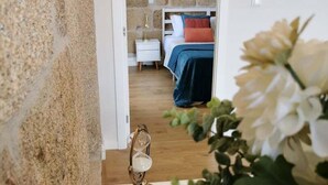 A doorway leading to a bedroom with a stone wall, a bed with a blue and red blanket, a bedside table with a lamp, and a wooden floor. - Solar dos Condes da Azenha (Sernancelhe)