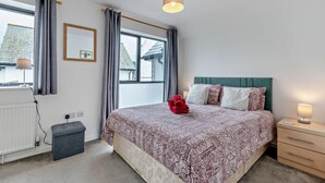 A bedroom with a bed, bedside tables, a window, curtains, a radiator, and a wall-mounted lamp. - 3 Bed in Ilfracombe (oc-s29585) (Ilfracombe)