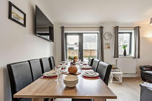 A dining area with a long wooden table, black cushioned chairs, and a view of the outside through large windows. - 3 Bed in Ilfracombe (oc-s29585) (Ilfracombe)