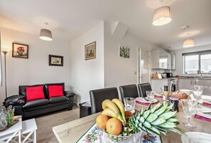 A modern living room with a dining area, featuring a black leather sofa, a wooden dining table with fruit and glassware, and a kitchen in the background.