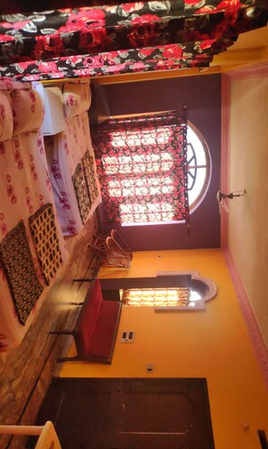 A compact room with a window, a bed with floral bedding, a small table, and a chair. - Hotel Sri Devi Comforts (Mudigere)