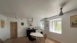 Eine moderne Küche mit einem weißen Tisch und Stühlen, einer Mikrowelle und einer Kaffeemaschine. Es gibt ein Fenster mit Blick auf Bäume und eine Tür mit Holzrahmen.