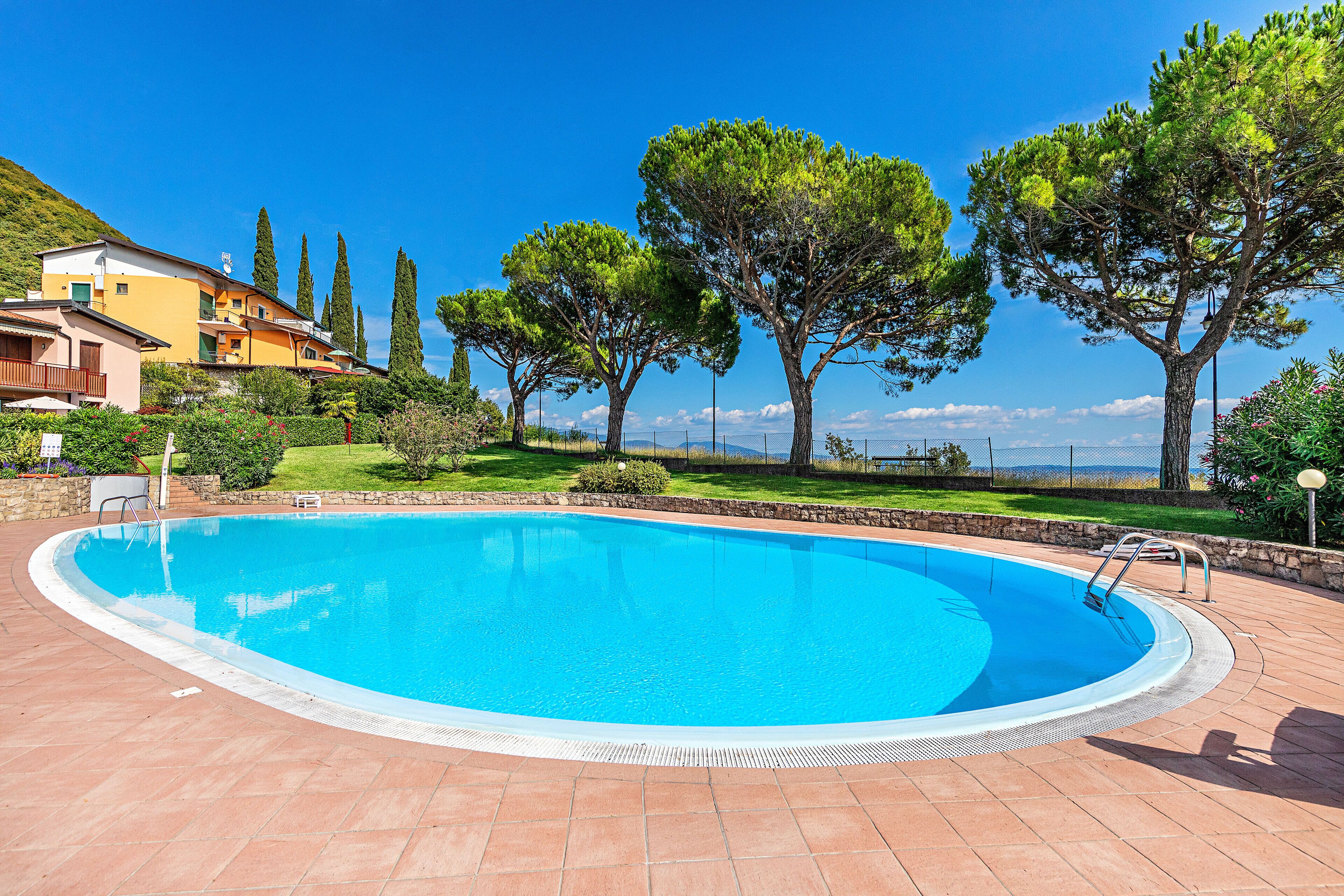 Une piscine dotée d’une terrasse carrelée, entourée d’arbres et bordée d’un bâtiment avec un balcon.