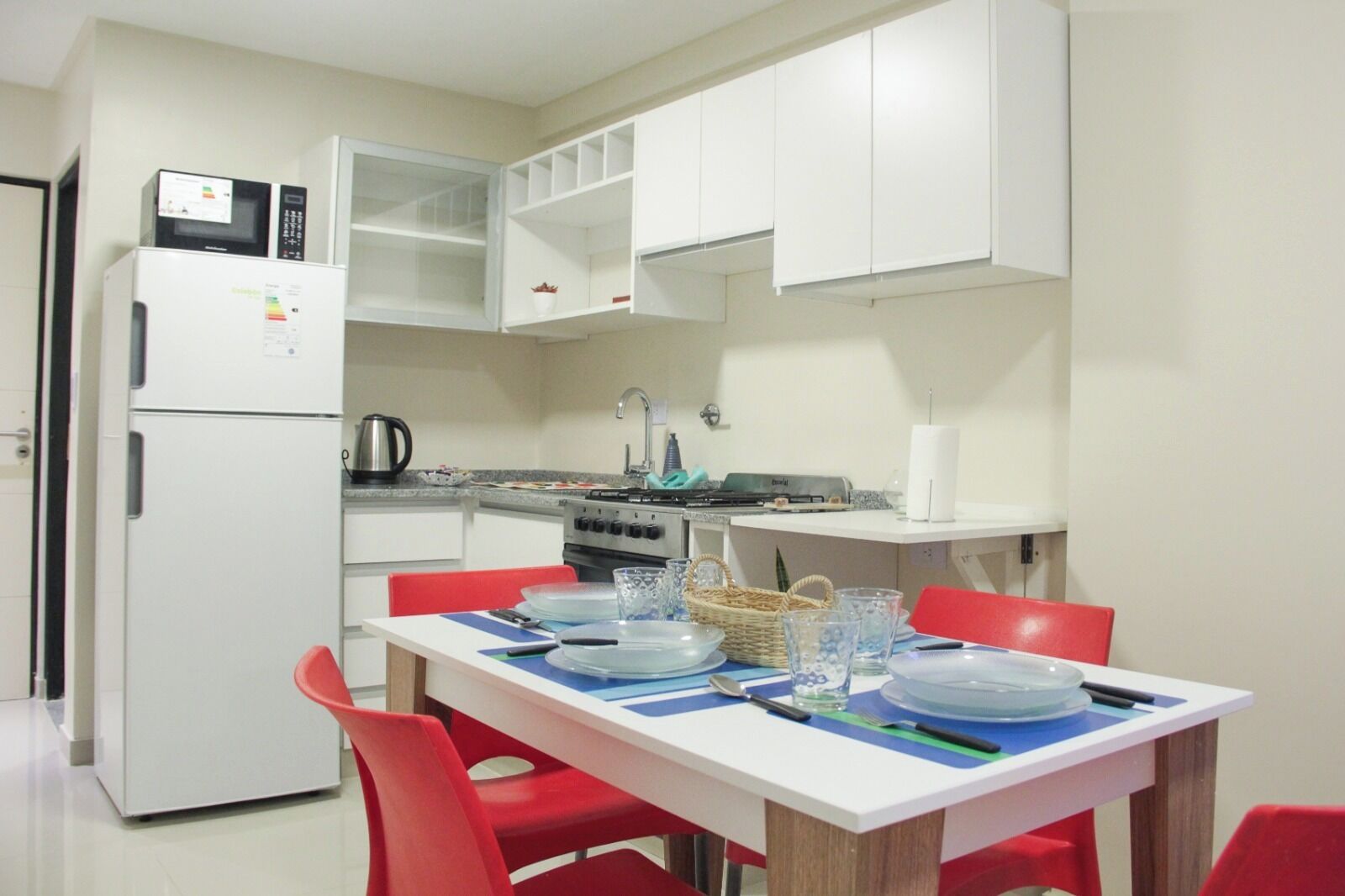 Una cocina compacta con armarios blancos, un refrigerador, una estufa y una mesa de comedor con sillas rojas.