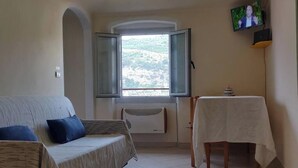 Una stanza con un divano, un tavolo con tovaglia, una finestra con persiane aperte e un televisore a parete.