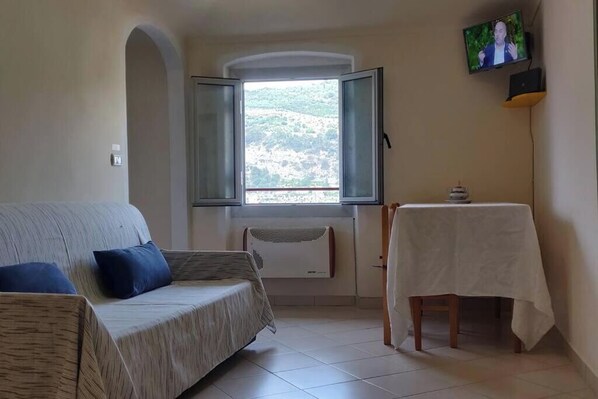 A room with a sofa, a table with a tablecloth, a window with open shutters, and a television on the wall.