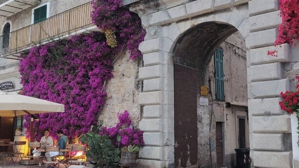 Un caffè all'aperto con tavoli e sedie, un edificio in pietra con un arco e fiori viola sulla parete.
