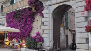 Un café al aire libre con mesas y sillas, un edificio de piedra con un arco y flores moradas en la pared.