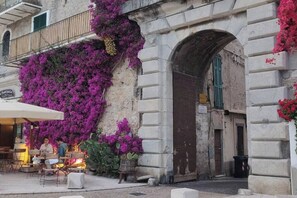 Un café en plein air avec des tables et des chaises, un bâtiment en pierre doté d’une arche, et des fleurs violettes qui ornent le mur.
