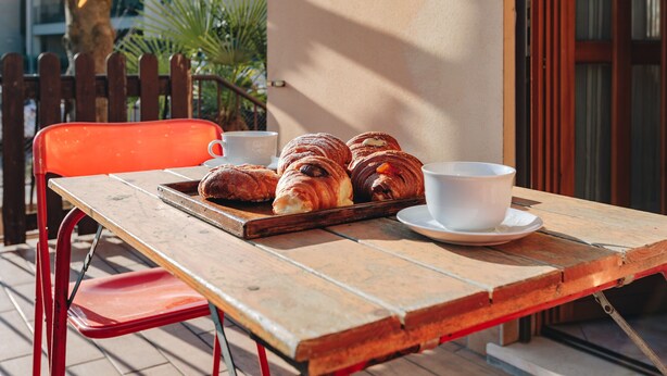 Una mesa de madera con una bandeja de croissants y una taza de café, una silla roja y una cerca de madera al fondo.