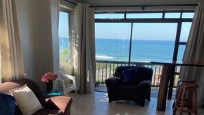 Een ruime woonkamer met een groot raam met uitzicht op de oceaan, een bruine bank, een donkere fauteuil en een kleine tafel met een bloevas.
