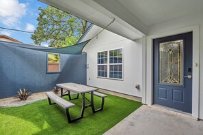 Une terrasse avec une table et des bancs, une porte ornée d’un panneau de verre décoratif et une petite fenêtre donnant sur un arbre.