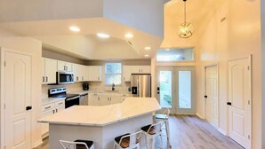 A modern kitchen with white cabinetry, a central island with bar stools, stainless steel appliances, and recessed ceiling lighting. - Seas the Day ¤ 3 mins to Florida #1 beach! (St. Augustine)