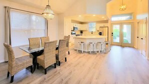 A modern kitchen with white cabinets, a central island, and a dining area with wicker chairs and a glass table. - Seas the Day ¤ 3 mins to Florida #1 beach! (St. Augustine)