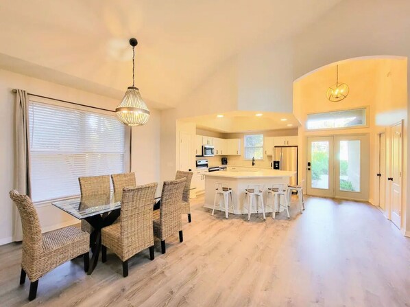 A modern kitchen with white cabinets, a central island, and a dining area with wicker chairs and a glass table. - Seas the Day ¤ 3 mins to Florida #1 beach! (St. Augustine)