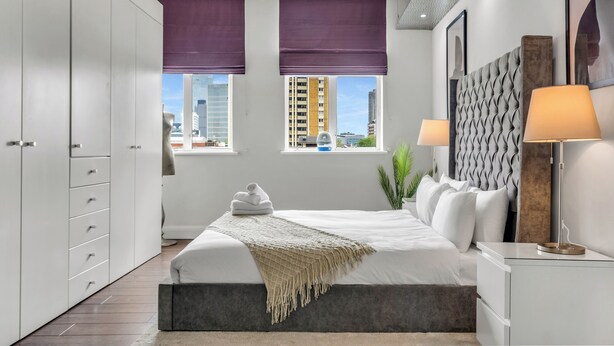 Une chambre moderne avec un lit, des tables de chevet, une armoire et de grandes fenêtres ornées de rideaux violets.