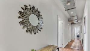 Un couloir avec un banc en bois, un miroir orné d’un motif en forme de soleil et un plafond laissant apparaître les conduits.