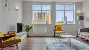 Un salon moderne doté d’une grande fenêtre, d’un téléviseur à écran plat, de chaises jaunes, d’un canapé gris et d’un tapis à motifs.