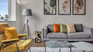 Un salon avec un canapé gris, un fauteuil jaune et un pouf à motifs. Trois reproductions botaniques encadrées ornent le mur.