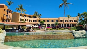 A hotel with a pool, palm trees, and a covered area with umbrellas.