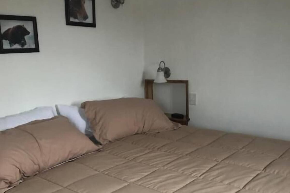 A bed with a beige quilt, a bedside table with a lamp, and two framed pictures on the wall.