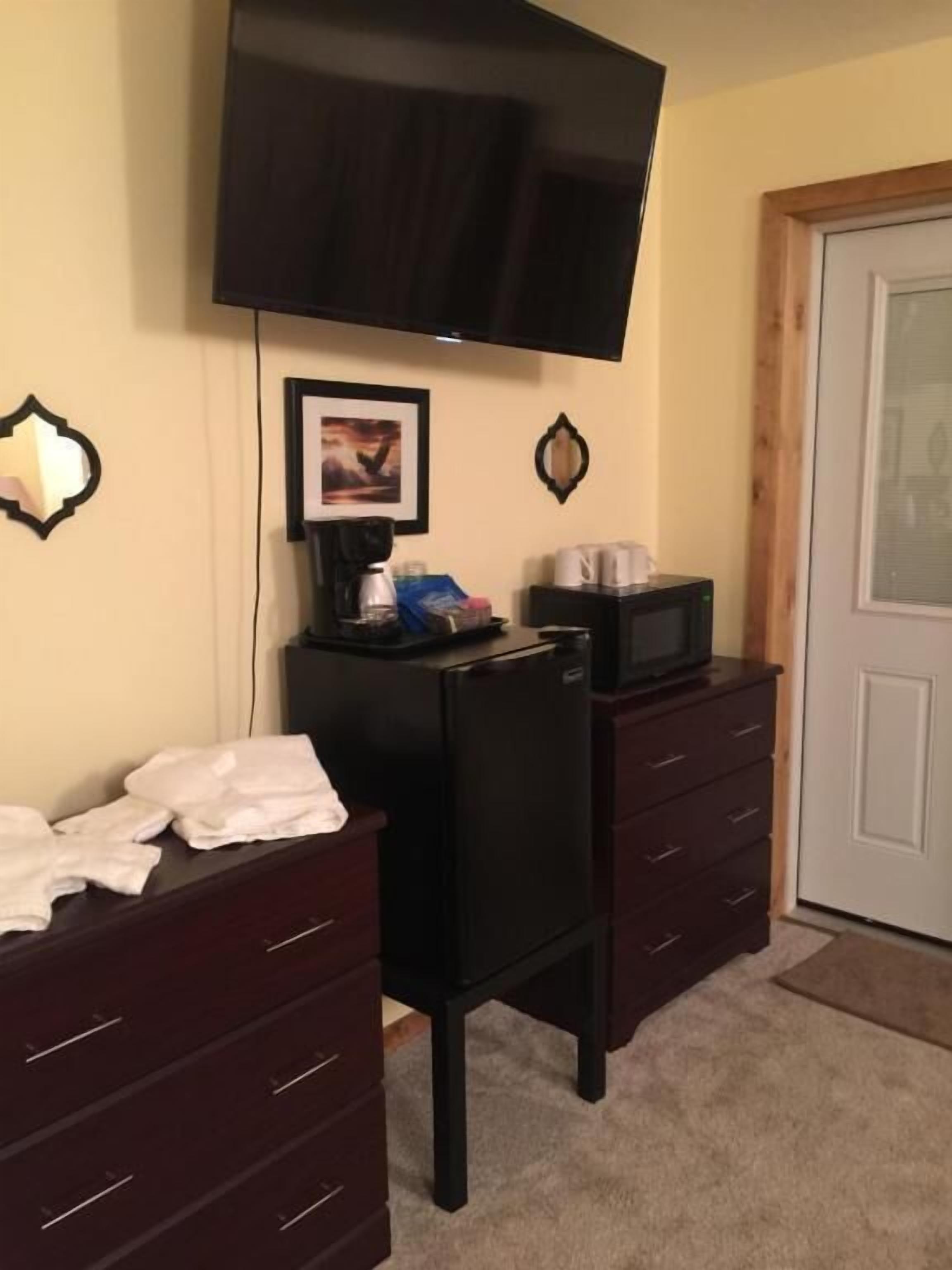 Quarto com televisão de tela plana na parede, pequeno espaço de cozinha com micro-ondas e cómoda de madeira escura.