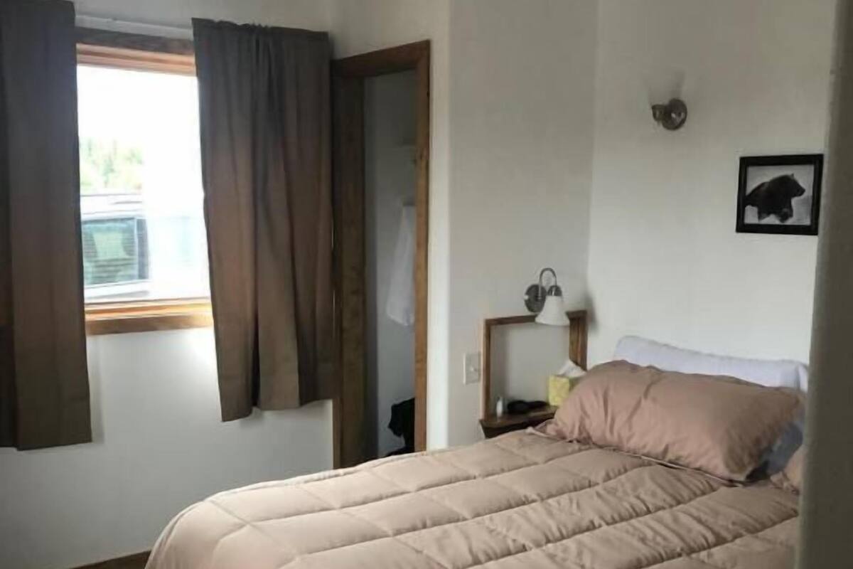 A bedroom with a large bed, a window with curtains, a bedside lamp, and a framed picture on the wall.