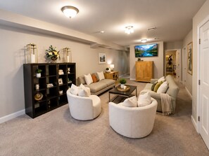 Ein Wohnzimmer mit einer Couch, zwei Sesseln, einem Couchtisch, einem Bücherregal und einem Fernseher.