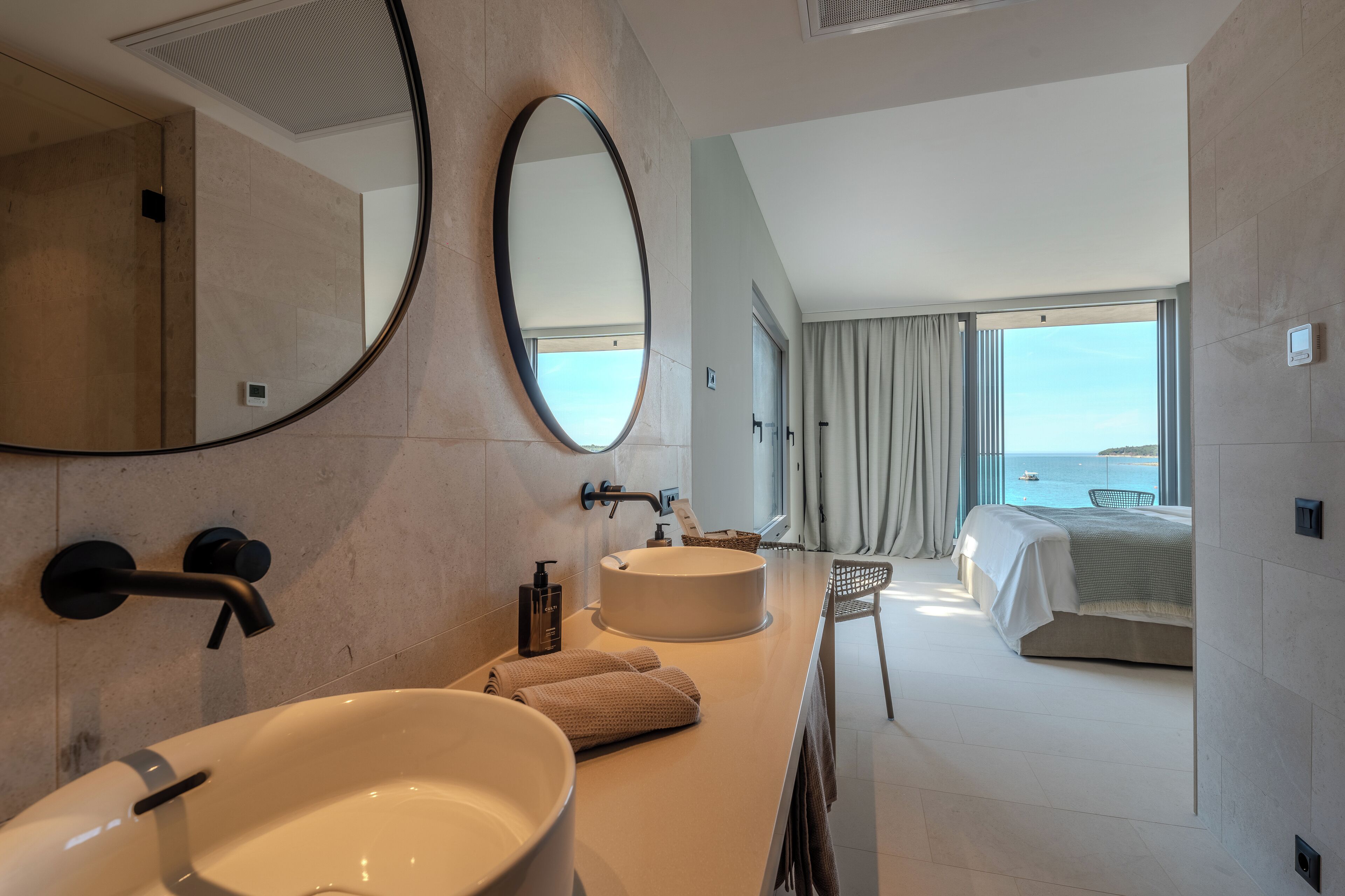 A modern bathroom with a large oval mirror, a round sink, and a bathtub. There is a bed with a view of the sea in the adjacent room.