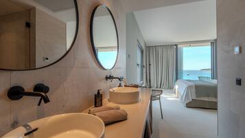 A modern bathroom with a large oval mirror, a round sink, and a bathtub. There is a bed with a view of the sea in the adjacent room.