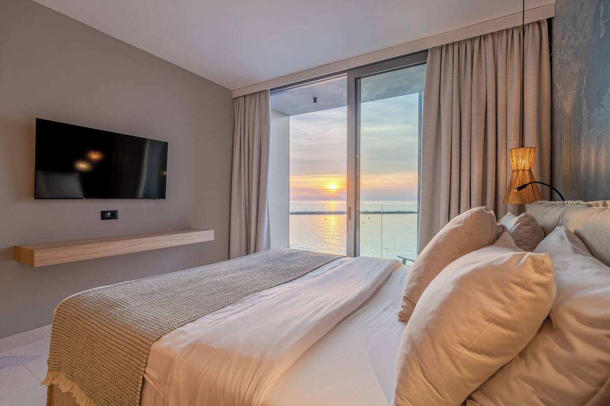 Ein Schlafzimmer mit Bett, Fernseher, Blick auf den Sonnenuntergang und einem großen Fenster.