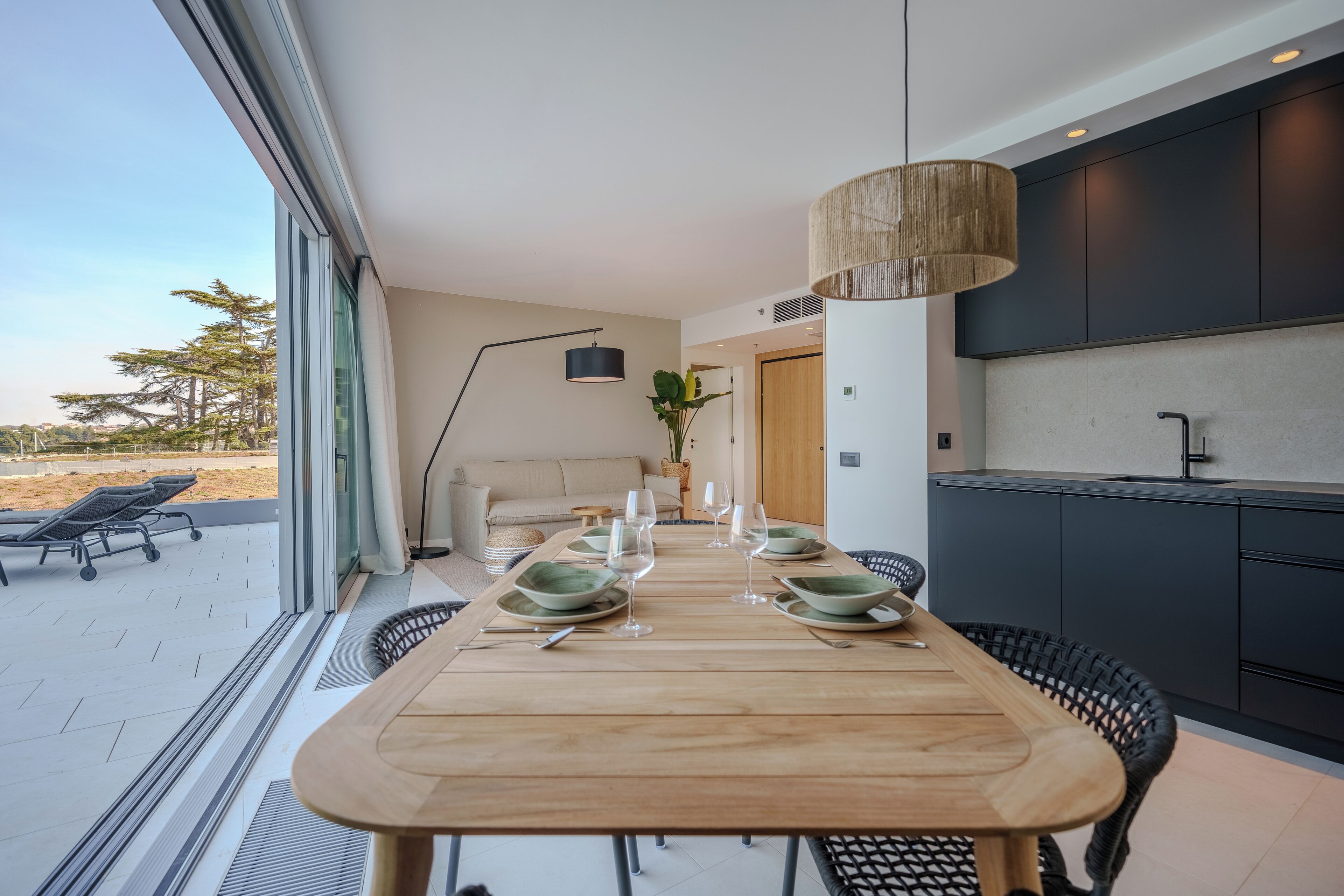 Ein moderner Essbereich mit einem Holztisch, gedeckt für vier Personen, eine Küche mit dunklen Schränken und Blick auf den Strand durch große Fenster.