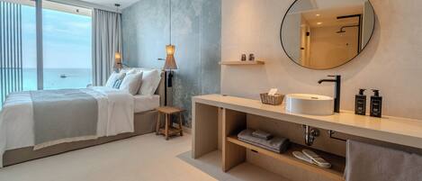 A modern hotel room with a large bed, a round mirror, a vanity with a sink, and a view of the sea.