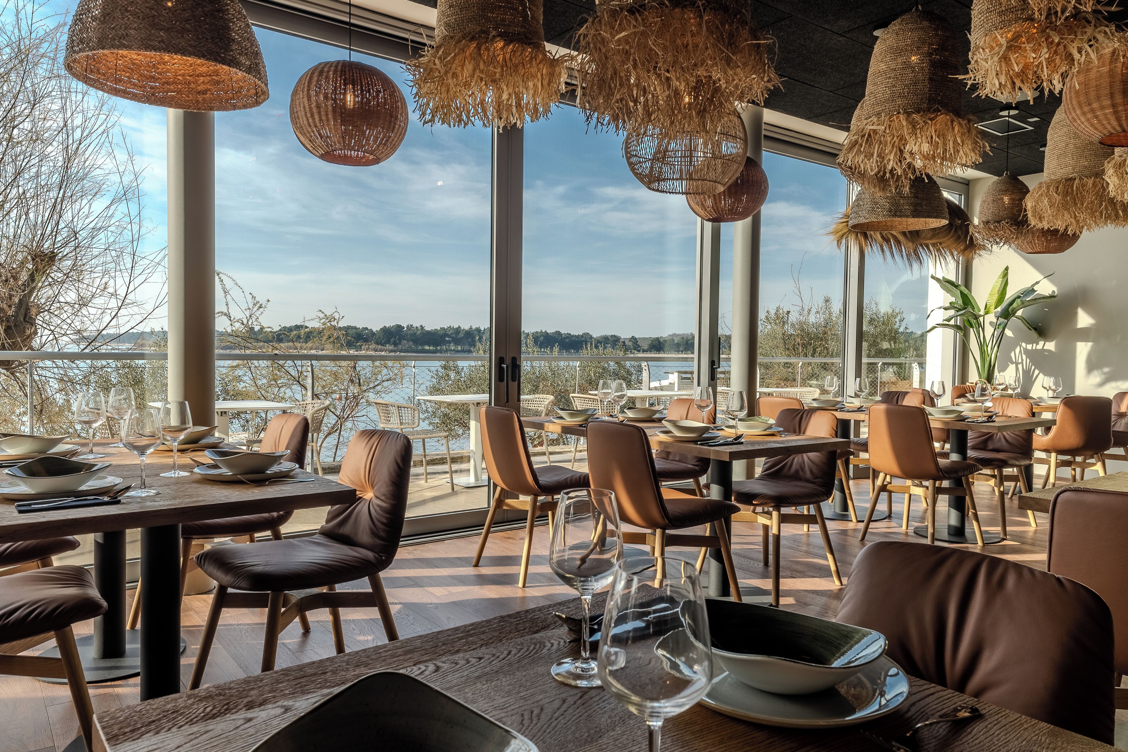 An outdoor dining area with tables and chairs, large windows overlooking a body of water, and hanging wicker lamps.