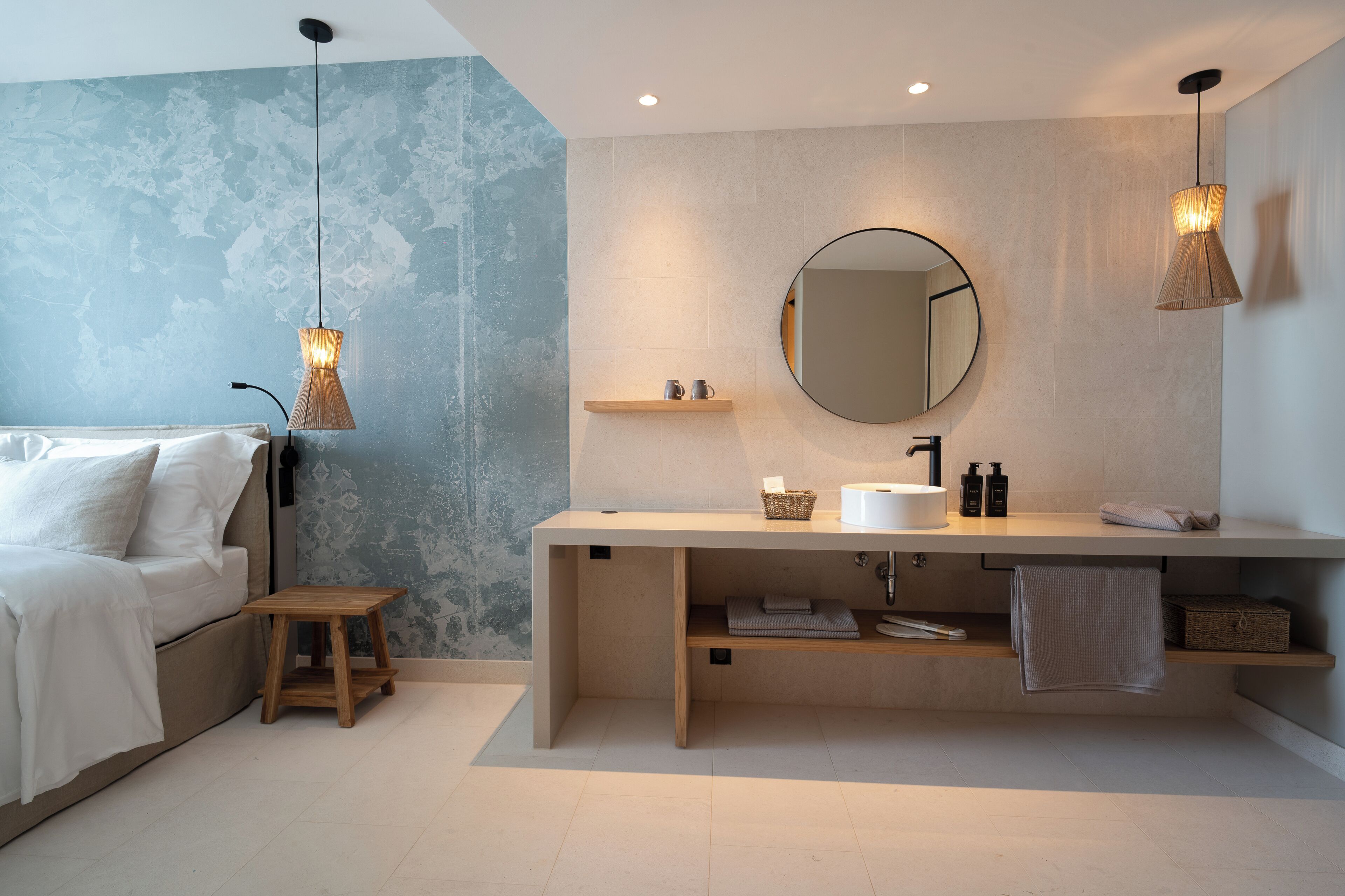 Un baño moderno con un espejo grande, un lavamanos y un estante de madera. En la habitación contigua, hay una cama con ropa de cama blanca.