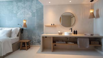 Un baño moderno con un espejo grande, un lavamanos y un estante de madera. En la habitación contigua, hay una cama con ropa de cama blanca.