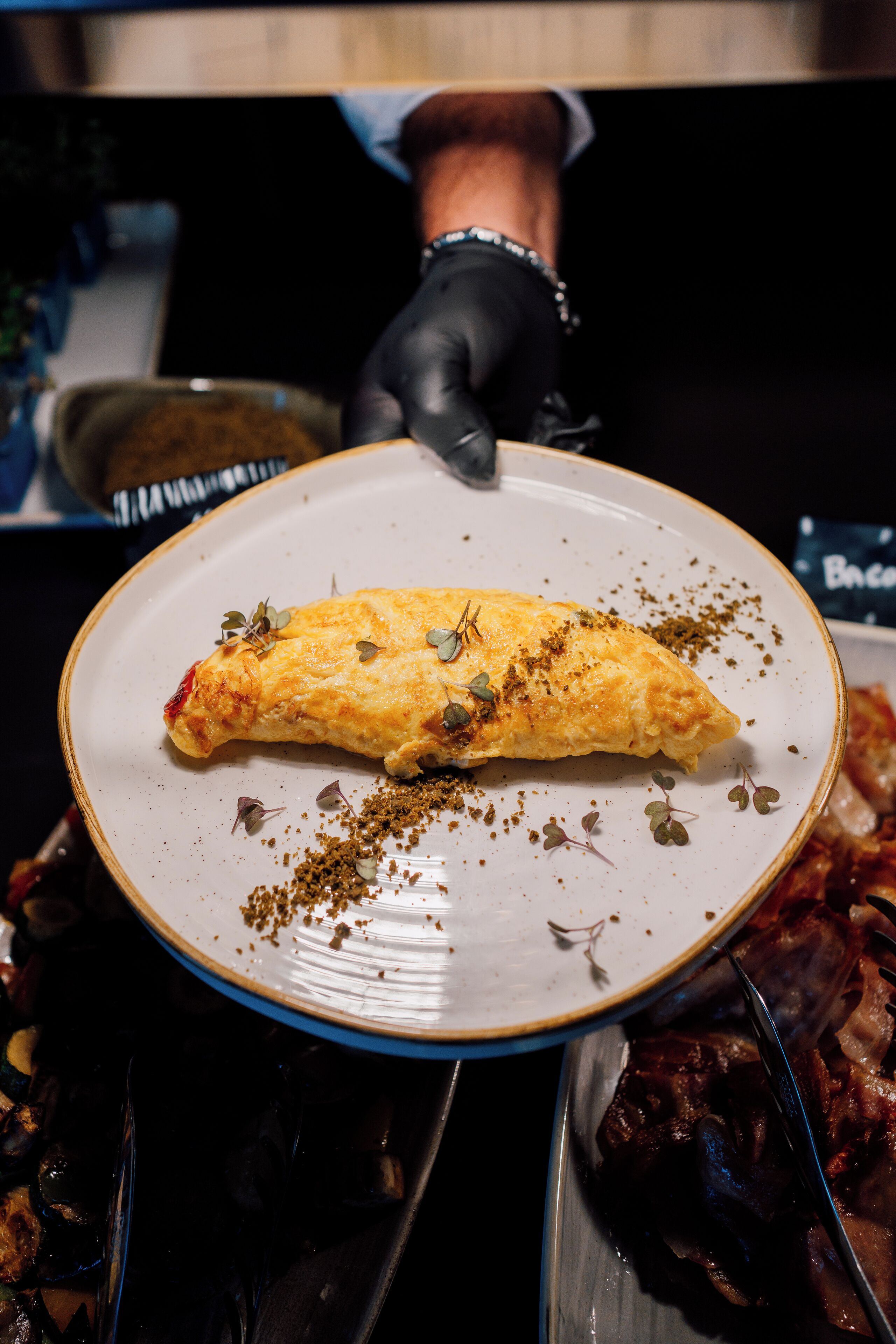 En tallerken med en sammenrullet omelet pyntet med krydderurter, holdt af en person med en sort handske.