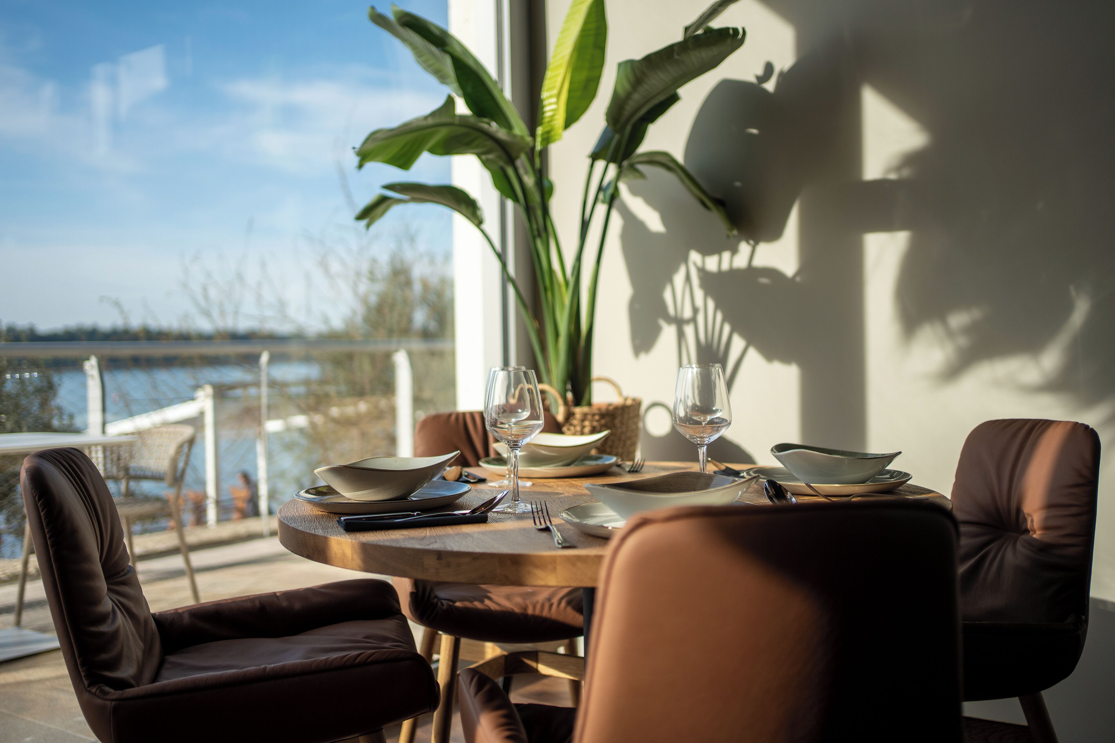A dining table set with plates, glasses, and cutlery, surrounded by leather chairs, with a view of the water outside.