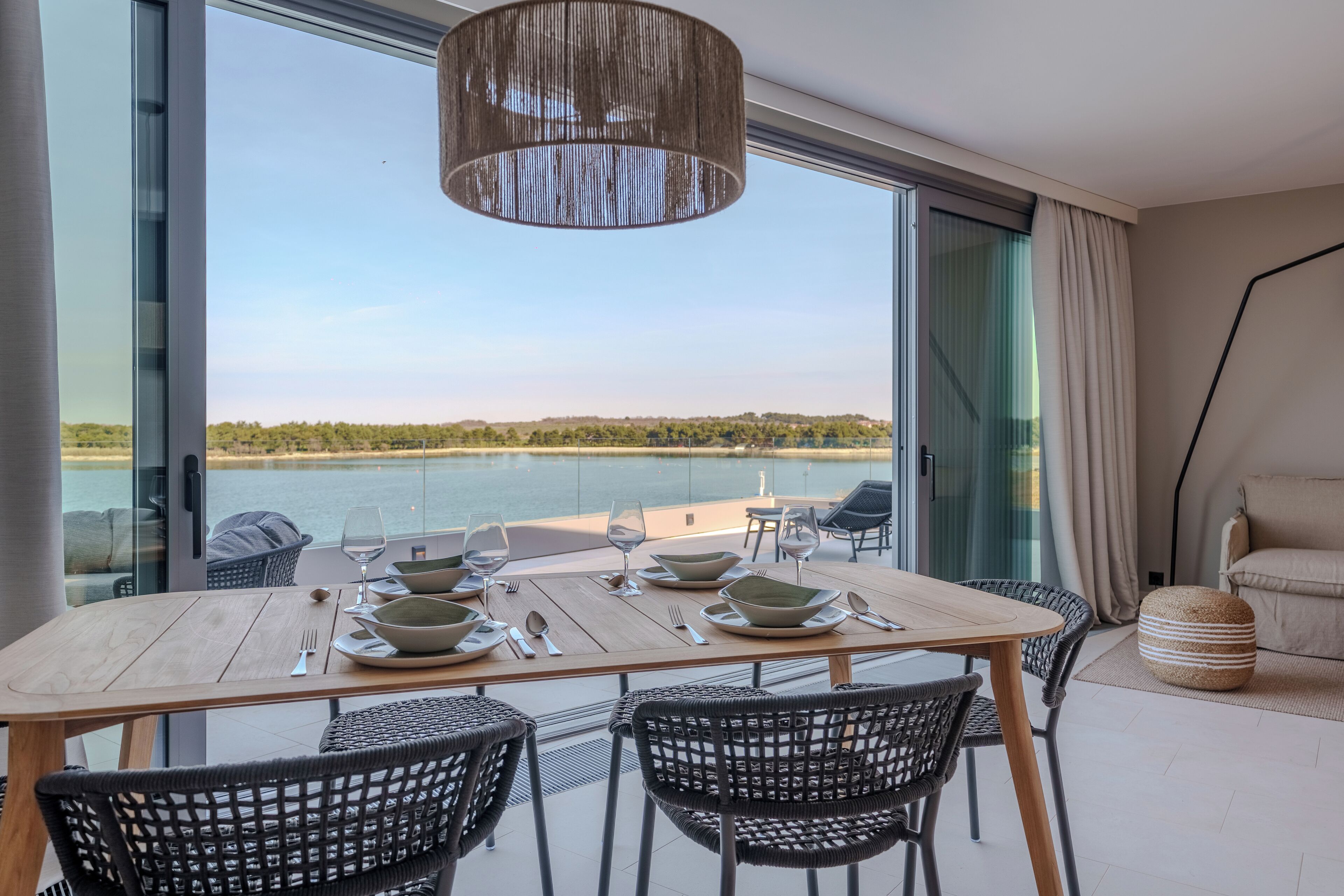 Comedor con mesa dispuesta para cuatro, vistas a un cuerpo de agua, amplia ventana y lámpara colgante moderna.