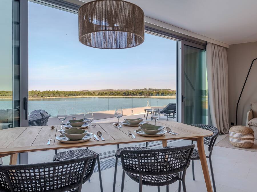 Comedor con mesa dispuesta para cuatro, vistas a un cuerpo de agua, amplia ventana y lámpara colgante moderna.