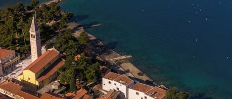 Vista aérea de un pueblo costero con una torre destacada, aguas azules cristalinas y un muelle largo que se adentra en el mar.