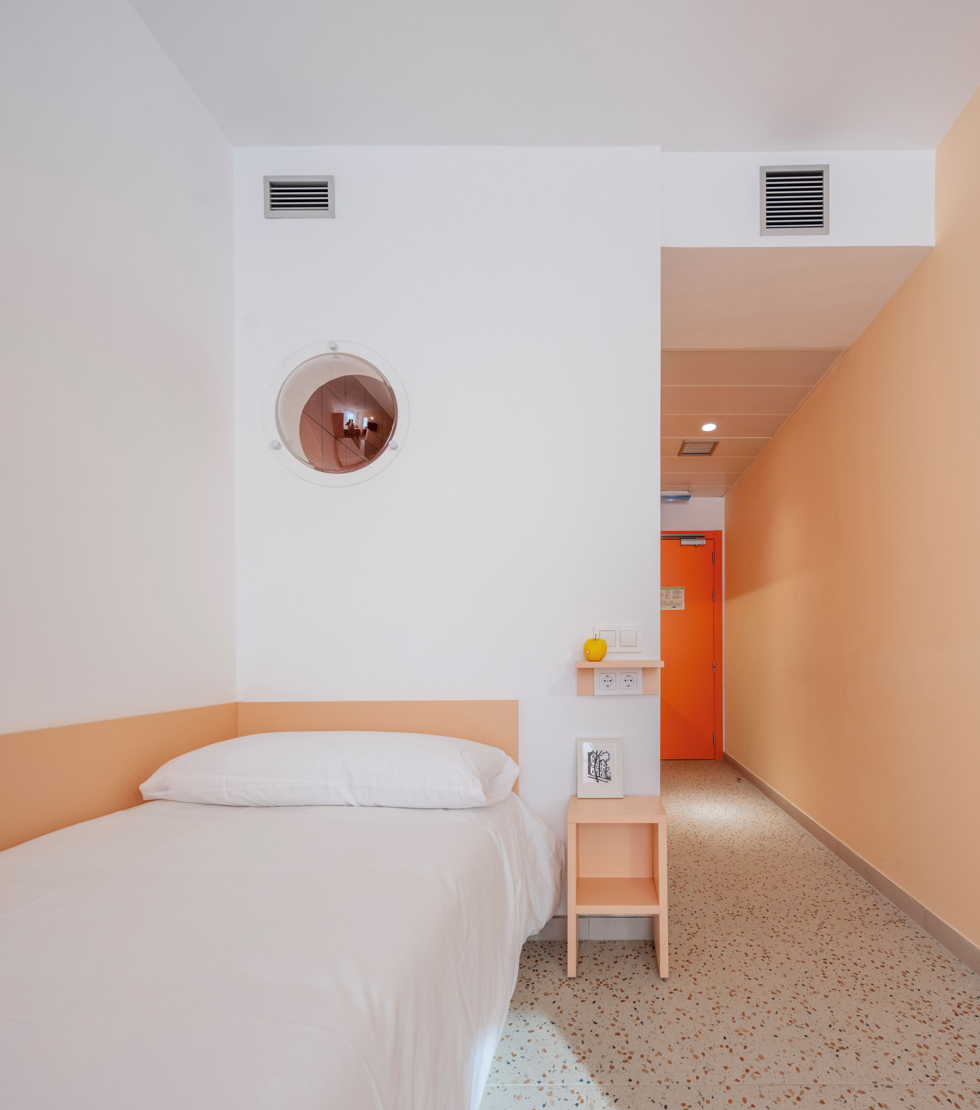 Una camera d'hotel con un letto, una piccola scrivania con una sedia, uno specchio rotondo e una porta che dà su un'altra stanza.