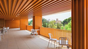 A covered outdoor area with wooden slats, white chairs, and tables, overlooking a green landscape with trees and buildings. -  Valencia Tower (Burjasot)