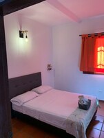 Uma cama de solteiro com cabeceira cinza, uma mesa de cabeceira pequena com um cesto e um luminário fixado na parede.
