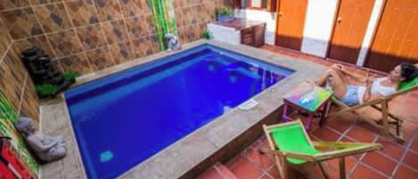 A small swimming pool with a tiled deck, a wooden chair, and a table.