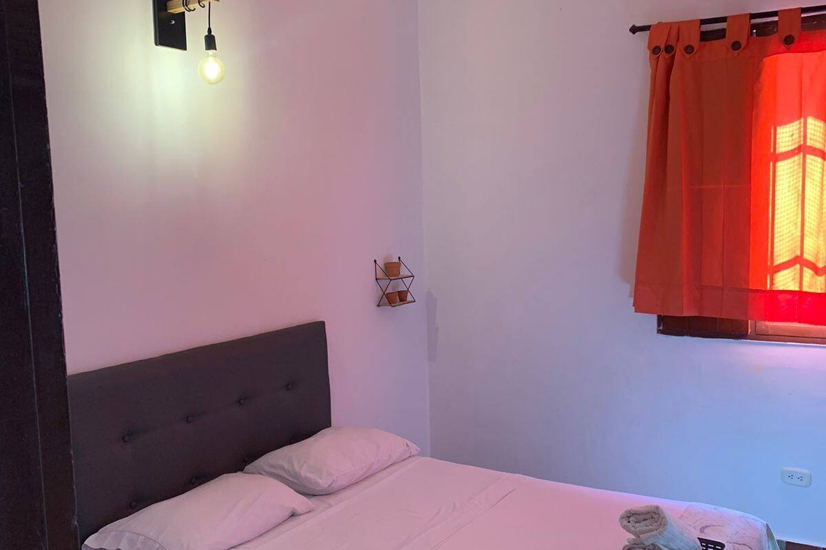 Un lit simple avec une tête de lit grise, une petite table de chevet avec un panier, et un luminaire fixé au mur.