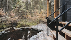 Mökki, jossa on puinen terassi, kivillä reunustettu lampi ja metsäinen tausta.