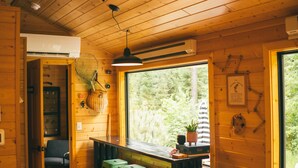 Interior acogedor de una cabaña con paredes de madera, un ventilador de techo, una zona de comedor con taburetes y un ventanal con vistas al exterior.