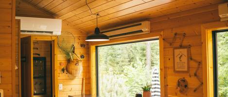 A cozy cabin interior with wooden walls, a ceiling fan, a dining area with stools, and a window offering a view of the outdoors.