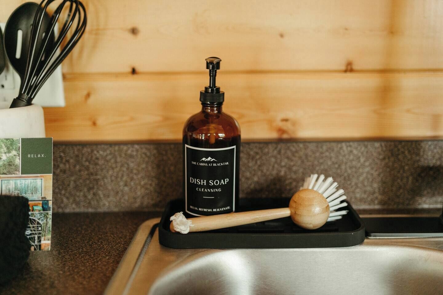 A bottle of dish soap on a black soap dish with a wooden brush beside it, placed on a kitchen counter.